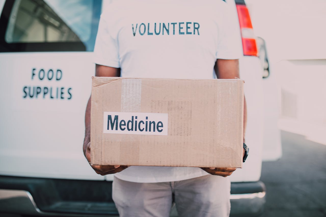 heros-img A volunteer holding a box labeled medicine near a delivery van marked food supplies.