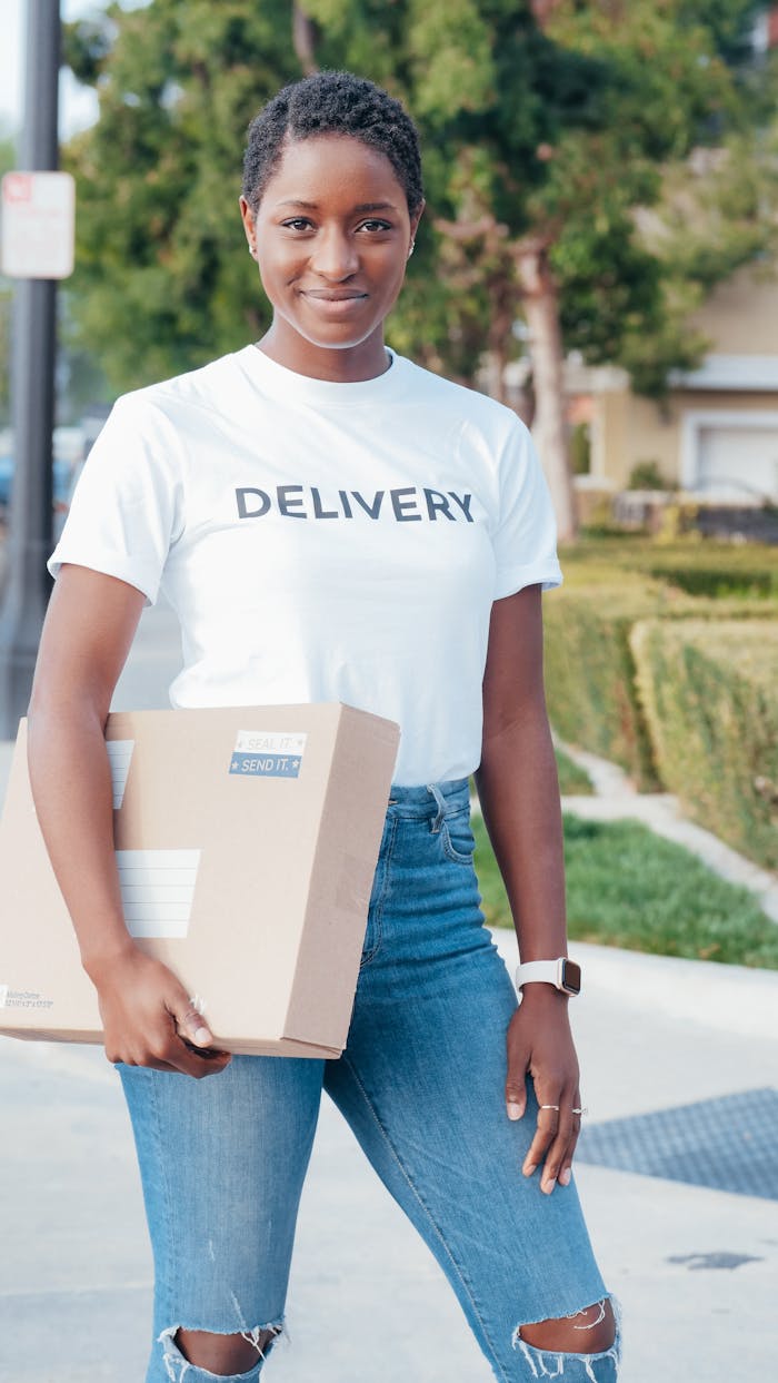 A young delivery woman smiles while holding a package outdoors, wearing a delivery t-shirt and jeans.